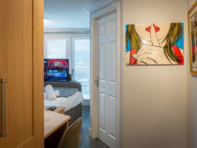 A hallway leading to a bedroom with a bed, TV, and desk with chair and a colorful artwork on the wall at Lakeland Lodge in Carnforth