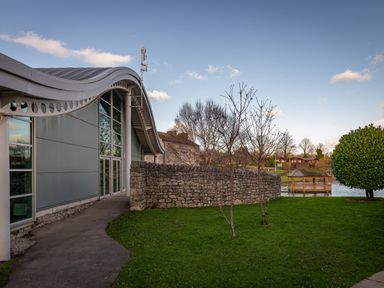A modern building with a curved roof beside a stone wall and small trees near a pond with a wooden gazebo at Lakeland Lodge in Carnforth