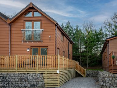 A brown wooden house with a balcony and a wooden fence next to a gravel pathway and trees at Lakeland Lodge in Carnforth