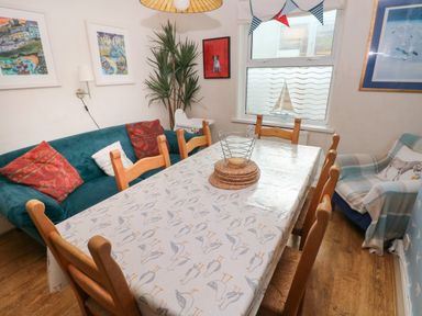 A dining room with a wooden table and six chairs a green sofa with pillows a plant high chair and window decorations at Greengate in Tenby
