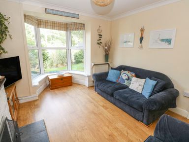 A living room with a blue sofa cushions a bay window with a wooden chest and framed pictures on the wall at Greengate in Tenby