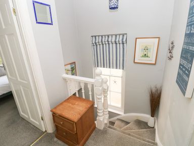 A staircase with carpet and a wooden chest of drawers at the top with framed pictures and a window with striped blinds at Greengate in Tenby