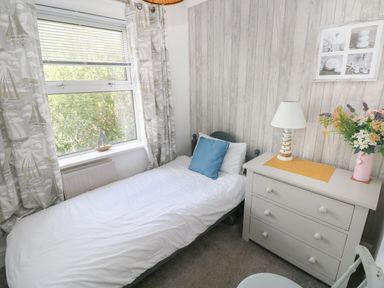 A small bedroom with a single bed, a chest of drawers with a lamp and a vase of flowers, and a window with patterned curtains at Greengate in Tenby