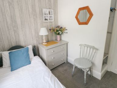 A bedroom corner with a single bed a blue pillow a small dresser with a lamp and flowers a grey chair and a hexagonal mirror on the wall at Greengate in Tenby