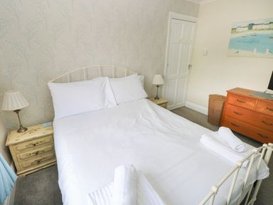 A bedroom with a bed with white linens and pillows bedside tables with lamps a wooden dresser a wicker laundry basket and a painting on the wall at Greengate in Tenby