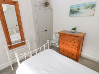 A bedroom with a white metal bed frame a wooden dresser a gray chair a gray wardrobe a large wooden framed mirror and a beach painting on the wall at Greengate in Tenby