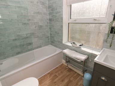 A bathroom with a bathtub, tiled walls, a window, a towel rack with towels, and a sink cabinet at Greengate in Tenby