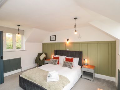 A bedroom with a double bed with pillows bedside tables a green chair and two windows at Rosemary Cottage in Briggswath near Sleights