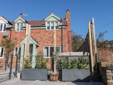 The front of a brick house with two dormer windows a porch two large plant pots with greenery and an outdoor seating area at Rosemary Cottage in Briggswath near Sleights