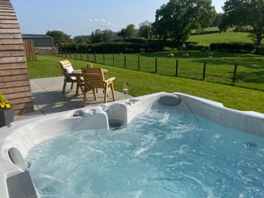 An outdoor hot tub with bubbling water next to a patio with wooden chairs and a table overlooking a fenced pasture with sheep at Y Dderwen in Bontuchel near Ruthin