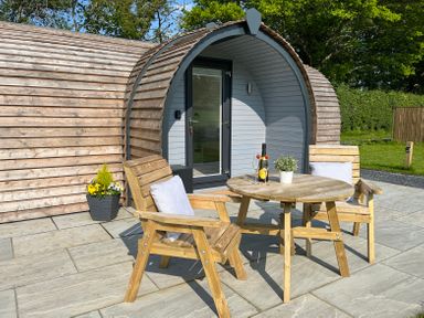 An outdoor seating area with wooden chairs and a round table in front of a curved wooden cabin at Y Dderwen in Bontuchel near Ruthin