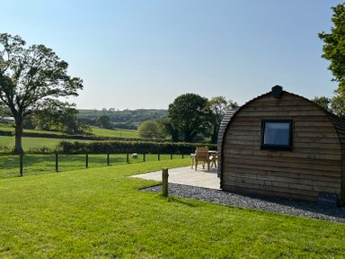 A curved wooden cabin with a window on grass near a patio with chairs and table in a rural landscape at Y Dderwen in Bontuchel near Ruthin