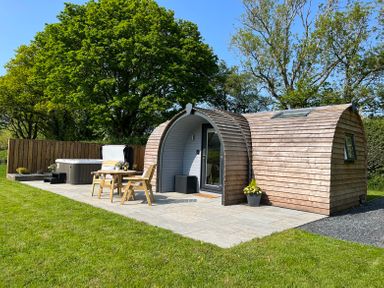 A wooden pod with outdoor chairs and table next to a hot tub on a stone patio with trees in the background at Y Dderwen in Bontuchel near Ruthin