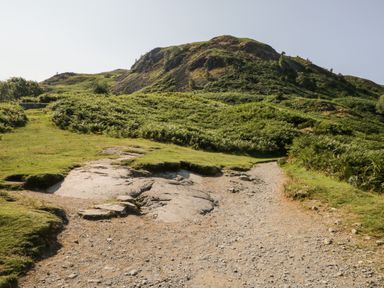 A dirt and gravel path leading towards a grassy hill with scattered bushes and rocks at 8 Oaks Field in Ambleside