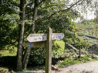 A wooden signpost on a dirt path with directional signs to Grasmere and Ambleside surrounded by trees and stone walls at 8 Oaks Field in Ambleside