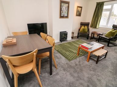 A living room with a dining table and chairs a television two armchairs a coffee table and a green rug at 8 Oaks Field in Ambleside