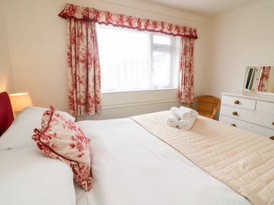 A bedroom with a bed with white and red floral pillow and beige quilt with rolled white towels on top white dresser with mirror and wicker chair by the window at 8 Oaks Field in Ambleside