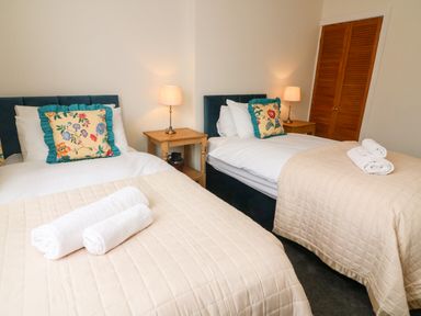 A bedroom with two single beds with beige quilts and floral pillows two bedside tables with lamps and rolled towels at 8 Oaks Field in Ambleside