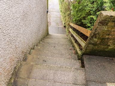 Outdoor concrete stairs between a textured wall and a hedge with a wooden railing leading to a paved walkway at 8 Oaks Field in Ambleside