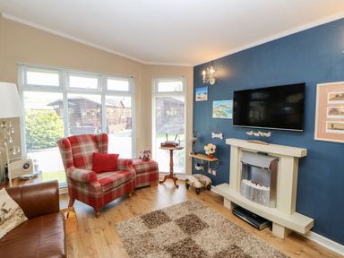 A living room with a plaid armchair, leather sofa, TV mounted above a fireplace, and large windows at 6 Badgers Retreat Tunstall near Richmond