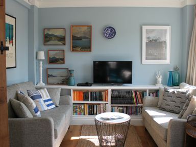 A sitting room with two sofas, a coffee table, a bookshelf with a TV on top, and paintings on the walls at 2 The Dunes Thorpeness