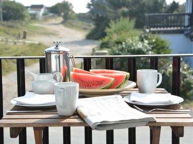 A wooden table on a balcony with two white mugs plates with napkins a pitcher a french press watermelon slices and a newspaper at 2 The Dunes Thorpeness