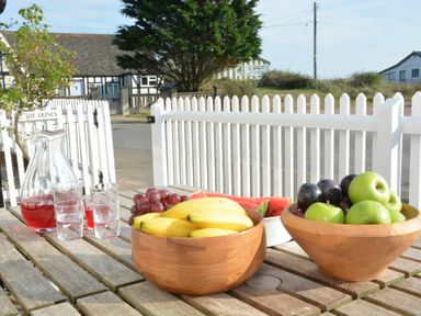 An outdoor wooden table with bowls of bananas grapes apples and a pitcher of red drink with glasses by a white picket fence at 2 The Dunes in Thorpeness