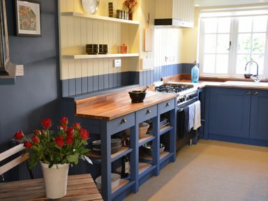 A kitchen with blue cabinets wooden countertops a gas stove a sink by a window and a pot of red roses on a wooden bench at 2 The Dunes Thorpeness