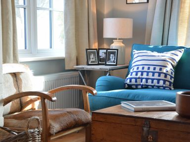 A living room with a blue sofa with a patterned pillow a wooden chair with animal print upholstery a side table with a lamp and black and white photos and a wooden chest as a coffee table with a book at 2 The Dunes Thorpeness
