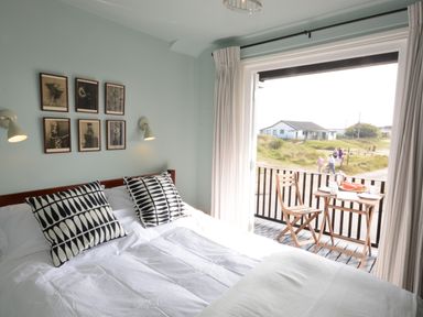 A bedroom with a bed with two patterned pillows wall lights framed pictures and a small balcony with a table and chairs at 2 The Dunes Thorpeness