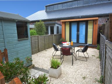 A small outdoor patio area with gravel ground a black metal table and three chairs a blue wooden shed and a modern wooden and glass building at Holmleigh Aldeburgh
