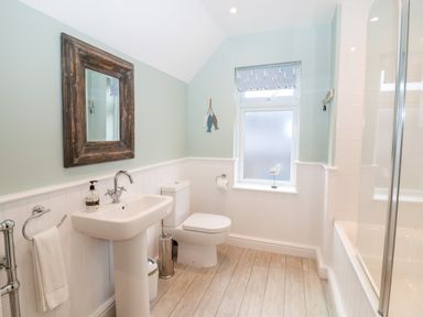 A bathroom with a pedestal sink, toilet, bathtub with glass screen, window with patterned blind, wooden framed mirror, and wooden floor at Holmleigh, Aldeburgh