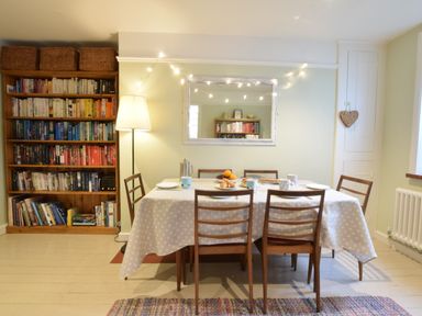 A dining room with a table covered by a polka dot tablecloth and six chairs a bookshelf with books and baskets a floor lamp a wall mirror with string lights and a heart decoration on the wall at Durdans Walberswick in Walberswick