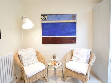 Two wicker chairs with cushions and a small table with coffee and food in a corner with a wall lamp and artwork at Durdans Walberswick