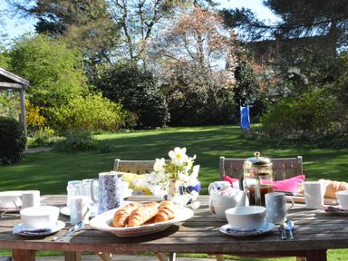 A wooden table set with croissants cups teapot and coffee maker in a garden with green grass and trees