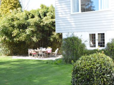 A patio with wooden chairs and a table surrounded by green bushes and a white house wall at Durdans Walberswick