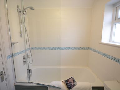A bathroom with a bathtub shower with glass screen white tiles and a book on a white towel at Bunny Cottage Aldeburgh