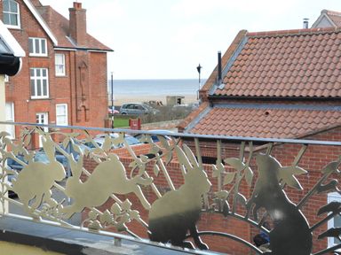 A metal balcony railing with cutouts of rabbits and foliage overlooking red brick buildings and a beach with the sea in the distance at Bunny Cottage Aldeburgh