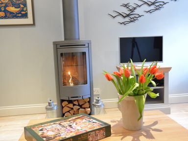 A living room with a wood burning stove a vase of orange tulips on a wooden table and a television on a stand at Bunny Cottage in Aldeburgh