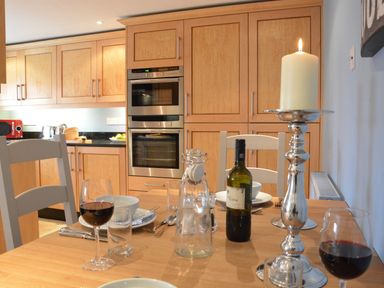 A kitchen with wooden cabinets a dining table set with wine glasses plates and a lit candle at Bunny Cottage Aldeburgh
