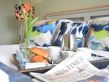 A breakfast tray with croissants cups a teapot a flower and a newspaper on a bed with blue and green pillows at Bunny Cottage Aldeburgh
