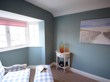 A corner of a room with a window and blue walls featuring a blue checkered armchair and ottoman a small white table with a glass bottle and a painting on the wall at Bunny Cottage Aldeburgh