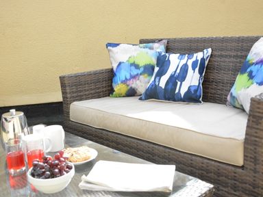 An outdoor seating area with a wicker sofa with cushions and a glass table with drinks grapes and cookies at Bunny Cottage Aldeburgh