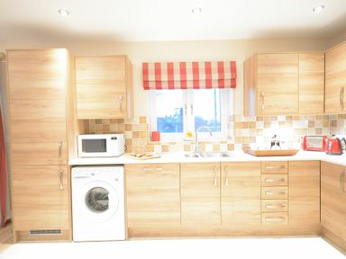 A kitchen with wooden cabinets a washing machine a microwave and a window with a red checkered blind at Red Robin Whepstead in Wickhambrook near Bury St Edmunds