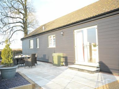 An outdoor patio area with dining table and chairs beside a building with black siding and glass double doors at Red Robin Whepstead in Wickhambrook near Bury St Edmunds
