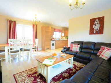 A living room with a black leather sofa and a wooden coffee table next to a dining table and wooden kitchen cabinets at Red Robin Whepstead in Wickhambrook near Bury St Edmunds