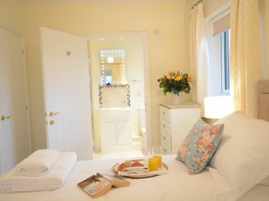 A bedroom with a bed with white bedding and a floral pillow a tray with croissants and orange juice towels a dresser with flowers a lamp and a view into a bathroom at Red Robin, Whepstead in Wickhambrook near Bury St Edmunds