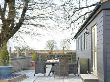 An outdoor patio with a table and four wicker chairs next to a building and trees in the background at Red Robin Whepstead in Wickhambrook near Bury St Edmunds
