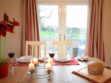 A dining table set with plates glasses lit candles and bread near glass doors with curtains at Red Robin Whepstead in Wickhambrook near Bury St Edmunds