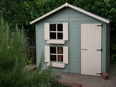 A green wooden garden shed with white windows and door surrounded by plants